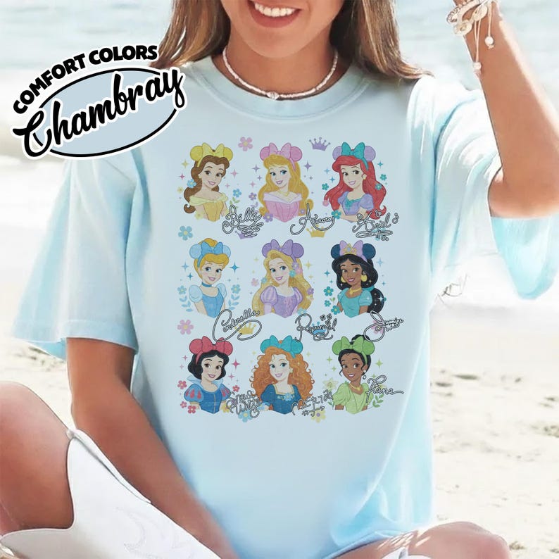 May include: Light blue t-shirt with a graphic of nine Disney princesses. The princesses are depicted with their names in cursive script. The shirt has the words "COMFORT COLORS" and "Chambray" printed on the upper left side.