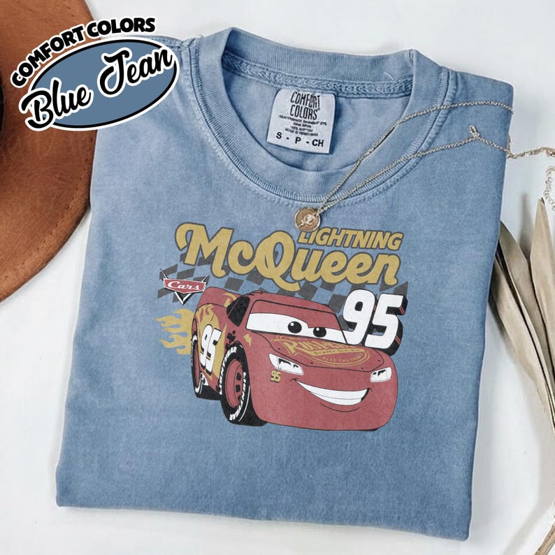 May include: Blue jean Comfort Colors t-shirt featuring a graphic of Lightning McQueen from the Cars movie. The design includes the number 95 and the text "Lightning McQueen". A silver necklace is draped across the shirt.
