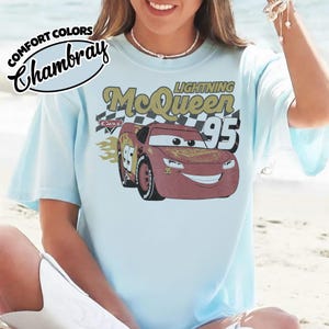 May include: Light blue t-shirt featuring a graphic of Lightning McQueen from the Cars movie. The design includes the text "Lightning McQueen 95" and a checkered flag. The shirt has short sleeves and a crew neck.