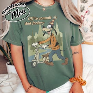 May include: Moss green t-shirt with a graphic of Goofy riding a scooter, wearing a party hat. The text "Off to commit dad foolery" is above the graphic. The shirt has the words "Comfort Colors" and "Moss" in a circular design.