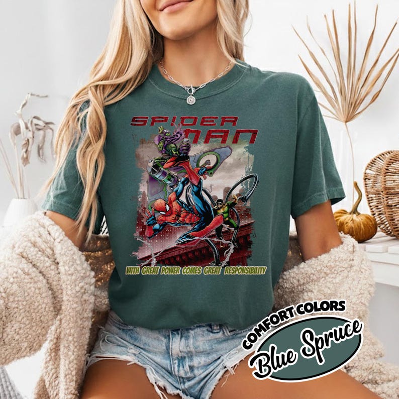 May include: Blue Spruce Comfort Colors t-shirt featuring a Spider-Man graphic. The design includes Spider-Man battling villains, with the text "SPIDER-MAN" and "WITH GREAT POWER COMES GREAT RESPONSIBILITY".