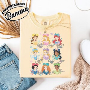 May include: A light yellow Comfort Colors sweatshirt featuring illustrations of Disney princesses. The princesses are arranged in two rows, with their names written below each image. The sweatshirt has the text "Comfort Colors Banana" in a circular design.