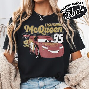 May include: Black t-shirt featuring a graphic of Lightning McQueen from the Cars movie. The design includes the text "Lightning McQueen 95" in gold and red, with a checkered background and the word "Cars". The shirt also has the words "Comfort Colors Black".