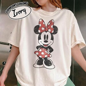 May include: Ivory-colored t-shirt featuring a distressed Minnie Mouse graphic. Minnie is depicted in black, white, and red, with a polka-dot bow and dress. The shirt has short sleeves and a crew neck. The words "COMFORT COLORS" and "Ivory" are printed on the top left.
