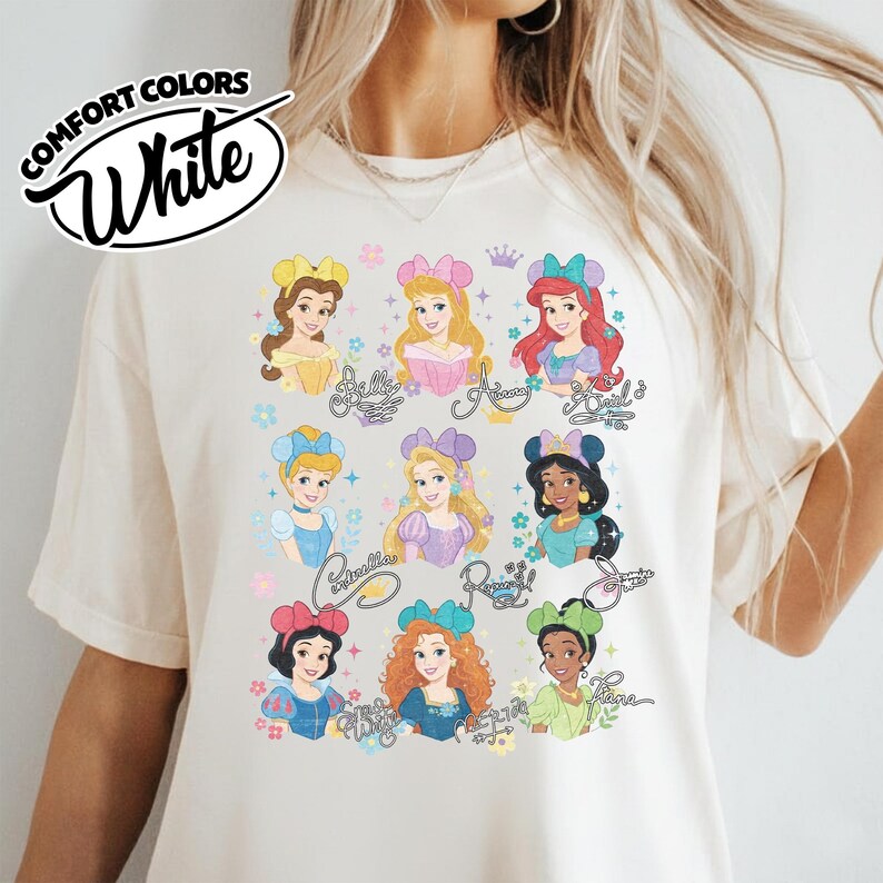 May include: White Comfort Colors t-shirt featuring a colorful graphic of Disney princesses. The design includes illustrations of Belle, Ariel, Cinderella, and others, with their names in cursive script. The shirt has a crew neck and short sleeves.