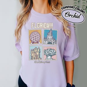 Może przedstawiać: Fioletowa koszulka z nadrukiem przedstawiającym cztery obrazy z Florydy w stylu znaczk&oacute;w pocztowych, w tym kulę, zamek i drzewo. Tekst "FLORIDA!!!" znajduje się nad obrazami, a "Walt Disney World" poniżej. Koszulka ma r&oacute;wnież tekst "COMFORT COLORS" i "Orchid".