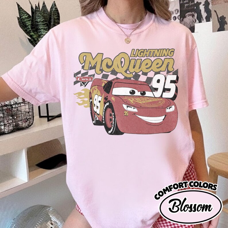 May include: Light pink t-shirt featuring a graphic of Lightning McQueen from the Cars movie. The design includes the text "Lightning McQueen 95" with a checkered flag background and flames. The shirt is labeled "Comfort Colors Blossom".