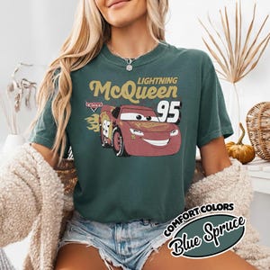 May include: Blue Spruce t-shirt featuring Lightning McQueen from the Cars movie. The design includes the text "Lightning McQueen 95" and a graphic of the character. The shirt is part of the Comfort Colors collection.