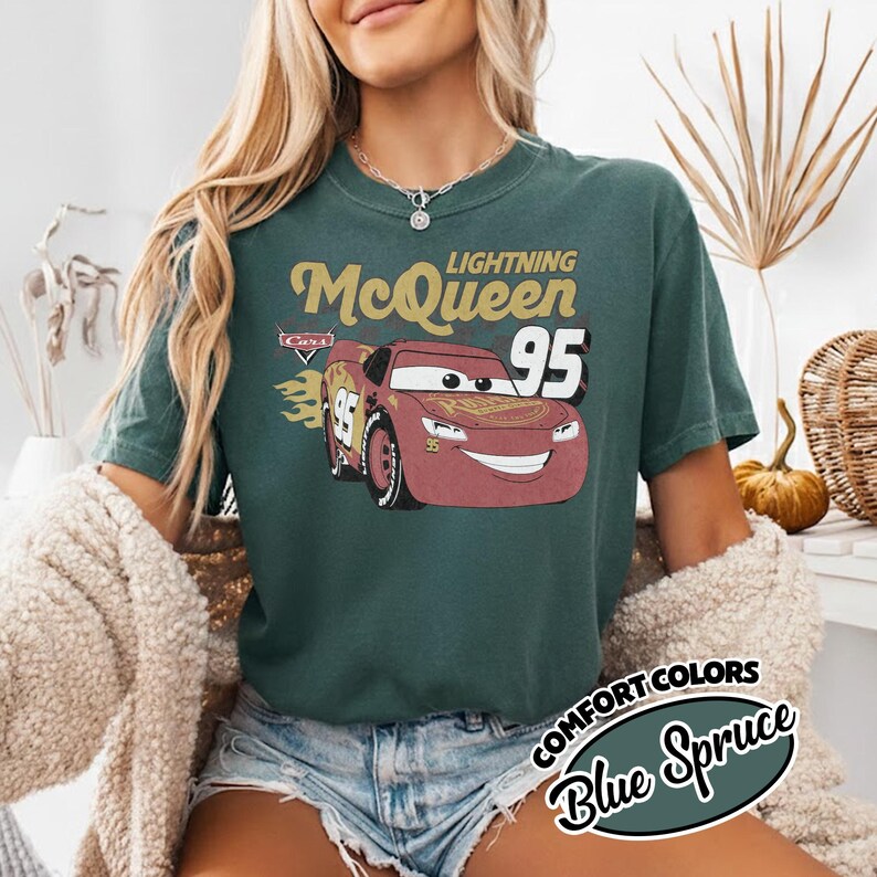 May include: Blue Spruce t-shirt featuring Lightning McQueen from the Cars movie. The design includes the text "Lightning McQueen 95" and a graphic of the character. The shirt is part of the Comfort Colors collection.