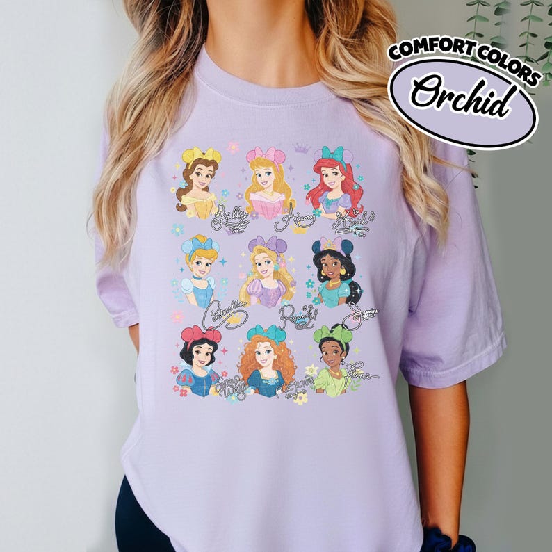 May include: A light purple t-shirt featuring illustrations of various princesses with their names in cursive. The shirt has the words "COMFORT COLORS" and "Orchid" printed on it. The shirt is a short-sleeved crew neck.