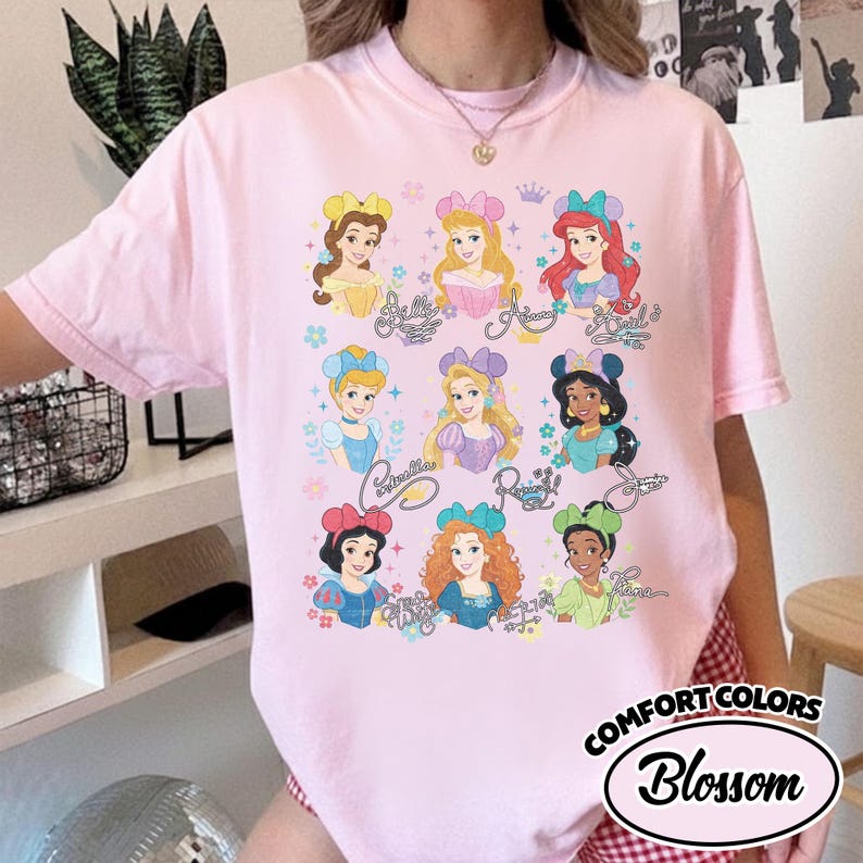 May include: A light pink Comfort Colors t-shirt featuring a graphic of Disney princesses. The princesses are depicted with their names in cursive script. The shirt has a round neckline and short sleeves. The text "Comfort Colors Blossom" is printed on the bottom right.