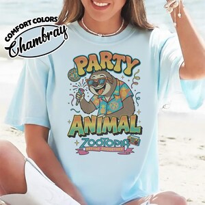 May include: Light blue t-shirt with the text "PARTY ANIMAL ZOOTOPIA" and a cartoon sloth wearing sunglasses and a Hawaiian shirt. The shirt also features a disco ball and a boombox. The words "Comfort Colors Chambray" are printed on the top left.