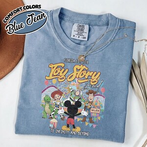 May include: A blue Comfort Colors t-shirt with a Toy Story Land design, showcasing characters such as Woody, Buzz Lightyear, and Mickey Mouse. The shirt includes the text "Florida Est. 2018" and "To Infinity and Beyond."