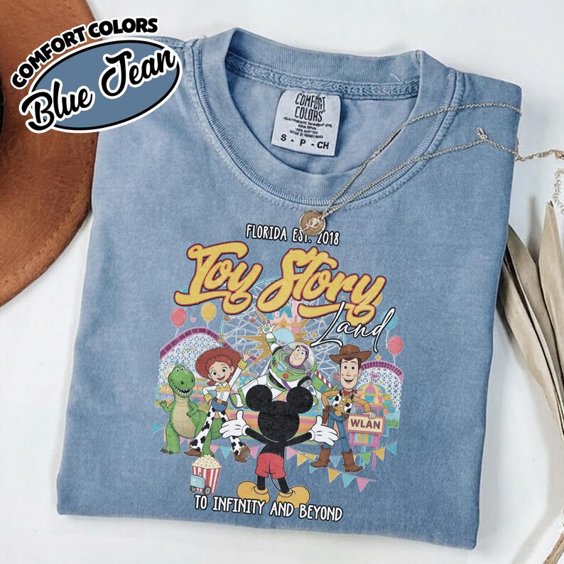 May include: A blue Comfort Colors t-shirt with a Toy Story Land design, showcasing characters such as Woody, Buzz Lightyear, and Mickey Mouse. The shirt includes the text "Florida Est. 2018" and "To Infinity and Beyond."