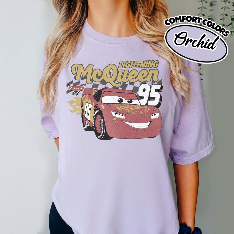 May include: A light purple t-shirt featuring a graphic of Lightning McQueen from the Cars movie. The design includes the text "Lightning McQueen 95" in gold and red, with a checkered flag background. The shirt is labeled "Comfort Colors Orchid".