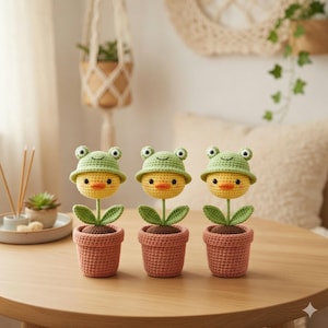 May include: Three handmade crochet flower decorations. Each features a yellow chick head wearing a green frog hat, green leaves, and a brown pot. The items are displayed on a wooden table.