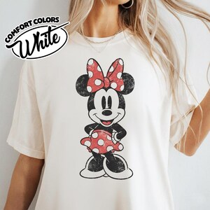 May include: White Comfort Colors t-shirt featuring a distressed Minnie Mouse graphic. Minnie is depicted in black, red, and white, wearing a polka dot bow and dress. The shirt has a vintage, worn look.