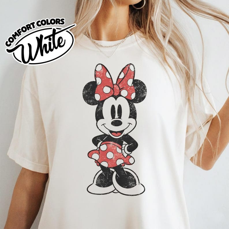 May include: White Comfort Colors t-shirt featuring a distressed Minnie Mouse graphic. Minnie is depicted in black, red, and white, wearing a polka dot bow and dress. The shirt has a vintage, worn look.