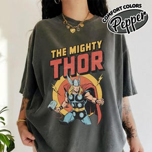 May include: Dark gray t-shirt featuring a comic book style illustration of Thor. The graphic includes the text "THE MIGHTY THOR" in red and yellow, with a drawing of Thor holding his hammer. The shirt has short sleeves and a relaxed fit.