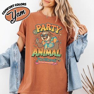 May include: A rust-colored t-shirt with a graphic of a sloth wearing sunglasses and a Hawaiian shirt, holding a microphone. The text reads "PARTY ANIMAL ZOOTOPIA" with additional text "BETTER ZOOGETHER". The shirt also has the text "COMFORT COLORS Yam".