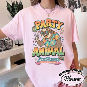 May include: Pink t-shirt with a colorful graphic featuring a sloth wearing sunglasses and a Hawaiian shirt, holding a microphone. The text reads "PARTY ANIMAL ZOOTOPIA BETTER ZOOGETHER". The shirt is from Comfort Colors, Blossom.