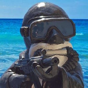 May include: A black figurine of a diver wearing a mask and snorkel, holding a small black submachine gun. The figure has a white beard and is dressed in a dark wetsuit, positioned against a backdrop of a bright blue ocean and clear sky.