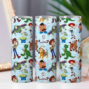 May include: Three cylindrical tumblers with a light blue background and a repeating pattern of cartoon characters. The characters include a cowboy, a space ranger, a green dinosaur, and other characters from the animated movie.
