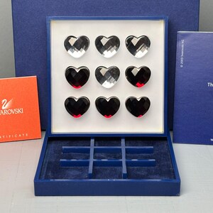 Peut inclure: Un jeu de morpion Swarovski composé de neuf cœurs en cristal facetté, transparents et noirs, disposés en grille dans une boîte de présentation bleue. L'ensemble comprend un coffret de rangement bleu avec un insert, un certificat et un livret bleu.