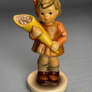 Pode incluir: Uma estatueta vintage de uma menina segurando um objeto amarelo em forma de cone. A menina usa um vestido marrom e mangas laranja, com sapatos e meias marrons. A estatueta está em uma base redonda de cor creme. O cone tem o número "18".