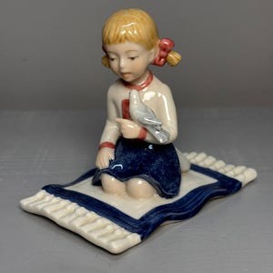 Op de afbeelding: Een porseleinen beeldje van een jong meisje dat op een klein kleedje knielt en een duif vasthoudt. Het meisje heeft blond haar met rode linten, draagt een wit shirt met rode bies en een blauwe rok. Het kleedje heeft een wit midden en een blauwe rand.