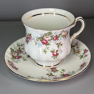 Puede incluir: Juego de taza y platillo blancos con un estampado floral de rosas rosas, rojas y azules. La taza y el platillo tienen ribetes dorados. La taza tiene un asa curvada.