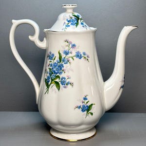 May include: A white ceramic teapot with a lid, decorated with blue floral patterns and green leaves. The teapot has a curved handle and spout, with gold trim. A classic piece for serving tea, perfect for a traditional tea setting.