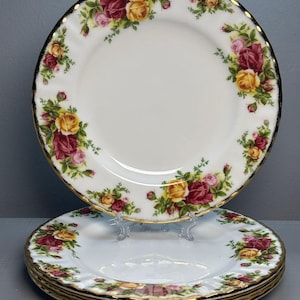 Puede incluir: Un juego de platos de porcelana blanca con un diseño floral de rosas en tonos rojos, amarillos y rosas. Los platos tienen un borde dorado y están apilados, con un plato ligeramente elevado. Diseño clásico.