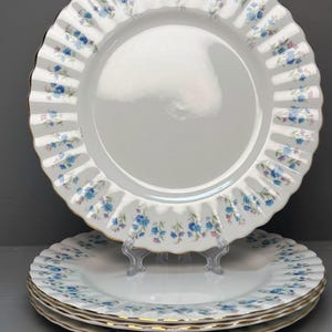 Peut inclure: Un ensemble de quatre assiettes en porcelaine blanche dotées d'un bord cannelé et festonné, orné d'un motif délicat de petites fleurs bleues et violettes. Chaque assiette présente un fin liseré doré sur le bord, offrant une finition raffinée.