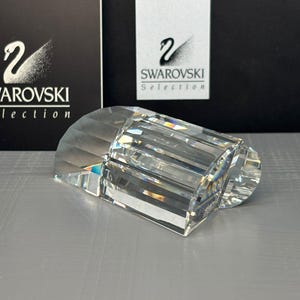 May include: A clear, faceted crystal paperweight with a rectangular, multi-sided design that refracts light. The object features sharp, geometric edges and a polished surface, displaying a small, etched Swarovski logo on one of its side facets.