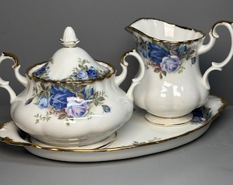May include: A white porcelain sugar bowl with a lid, a creamer, and an oval tray, all featuring a blue and lavender rose floral pattern with gold trim. The sugar bowl has two handles, and the creamer has a single handle and a scalloped rim.