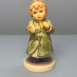 Könnte beinhalten: Eine Keramikfigur eines Kindes mit grünem Mantel, gelbem Schal und beiger Kapuze. Das Kind hat blondes Haar, dunkle Augen und steht auf einem runden Sockel. Der Mantel ist mit drei dunklen Knöpfen auf der Vorderseite und Taschen versehen.