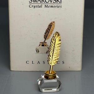 Peut inclure: Un objet de collection Swarovski Crystal Memories avec une plume dorée dans un encrier en cristal transparent. L'encrier a un design à facettes. La boîte porte le logo Swarovski et les mots "Crystal Memories".