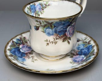 May include: A white teacup and saucer set decorated with blue and purple rose floral patterns. The rims are edged with gold. The teacup has a curved handle with gold detailing. The saucer is round with a scalloped edge.