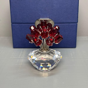Pode incluir: Um vaso de cristal com um buquê de flores vermelhas. O vaso tem formato de diamante e está em frente a uma caixa azul. As flores são feitas de cristais vermelhos.