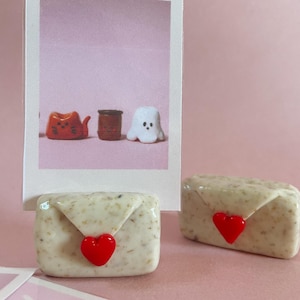 Peut inclure: Deux petits objets beiges en forme d'enveloppe avec une fermeture en forme de cœur rouge sont au premier plan. Une photo de style polaroid en arrière-plan montre un chat, un tonneau et un fantôme. Le fond est rose uni.