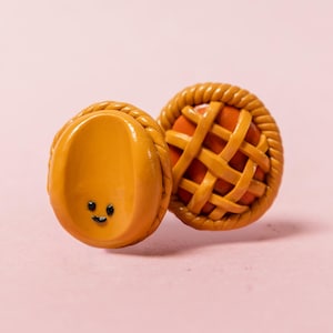 Op de afbeelding: Twee miniatuur oorbellen in de vorm van een taart. Eén oorbel heeft een lachend gezicht, de andere een roosterpatroon. Beide zijn oranje met een gevlochten rand, tegen een roze achtergrond. Ongeveer 2 cm in diameter.