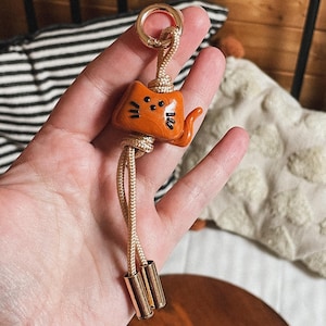 Peut inclure: Porte-clés en forme de chat orange avec des détails noirs, un fermoir doré et un cordon doré. Le porte-clés a une petite queue et des moustaches. Le cordon se termine par deux perles cylindriques dorées.