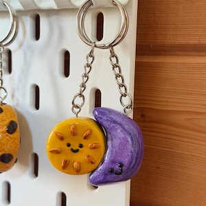 Può includere: Un portachiavi fatto a mano con un ciondolo a forma di sole e luna sorridenti. Il sole è giallo con raggi arancioni e la luna è viola con una finitura glitterata. I ciondoli sono collegati da una catena d'argento.