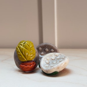 Puede incluir: Tres objetos decorativos de piedra. Uno presenta un diseño de hoja amarilla sobre una maceta roja. Otro es una piedra gris con pequeñas hendiduras redondas. El tercero es una piedra blanca con un patrón de pequeños círculos.