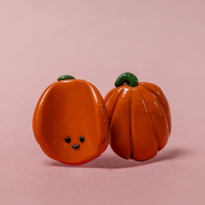 Könnte beinhalten: Zwei orangefarbene Kürbis-Ornamente mit grünen Stielen. Einer der Kürbisse hat ein lächelndes Gesicht. Die Kürbisse sind vor einem rosa Hintergrund platziert. Diese Dekorationsartikel sind wahrscheinlich für den Herbst oder Halloween.