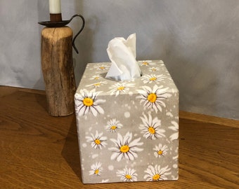 May include: A square tissue box cover with a white background and a pattern of white daisies with yellow centers. The box is made of wood and has a slot for tissues.