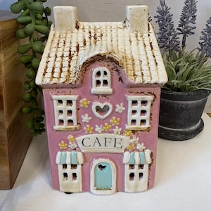 May include: Pink ceramic house ornament with a white roof and chimney. The house features windows, a heart-shaped cutout, and the word "CAFE" above the door. Decorative flowers and awnings add detail. A charming accent piece for home decor.