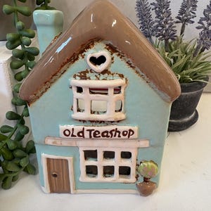 Puede incluir: Una casa de cerámica turquesa con techo marrón y el texto "OLD TEASHOP". La casa tiene un recorte en forma de corazón y una pequeña chimenea. Una pequeña planta en maceta está al lado de la casa.