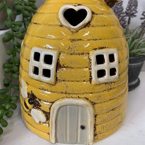 Könnte beinhalten: Ein gelbes Keramik-Bienenstockhaus mit einem herzförmigen Fenster, einer kleinen Tür und zwei Fenstern. An der Seite des Hauses befindet sich eine Biene.